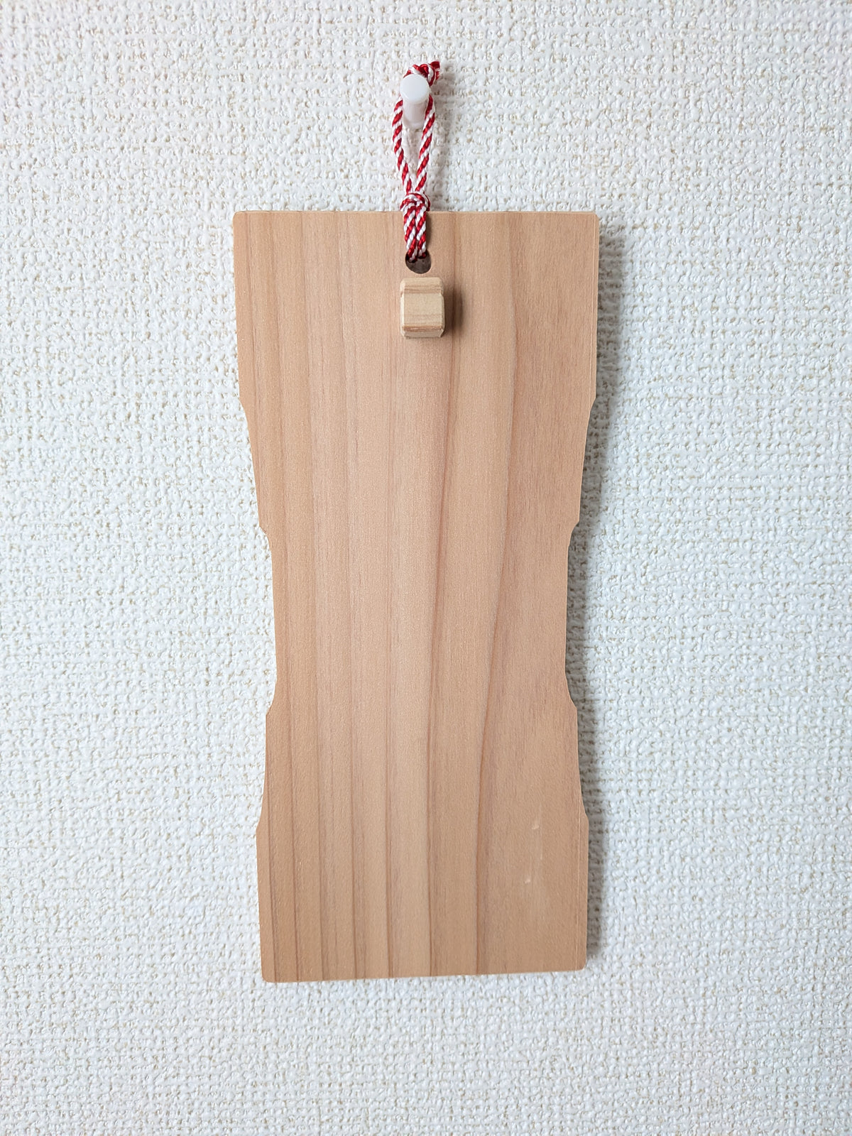 inori 壁掛けver.を壁に掛け、正面から見たところ。中ほどがくびれた杉の板で、上部に紅白の紐と小さな木の突起がついている